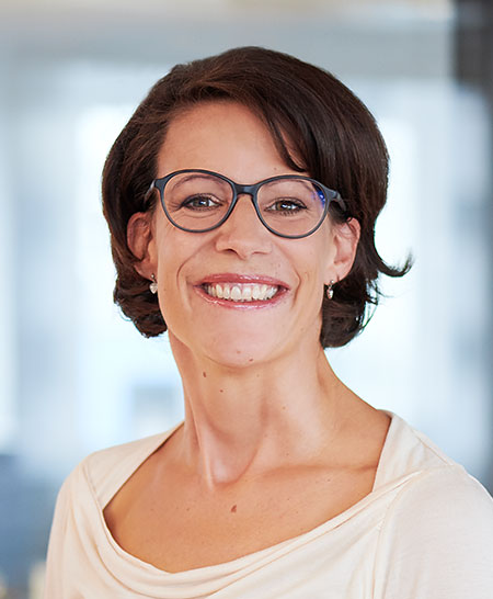 Eine lächelnde Frau mit Brille, braunen Haaren und einem cremefarbenen Oberteil.