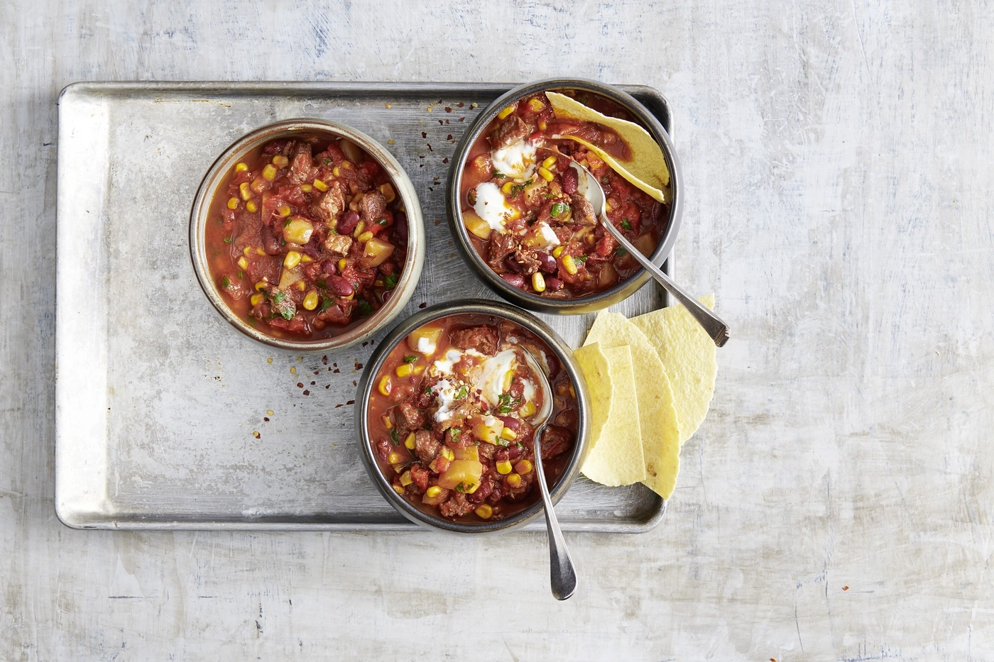 Trois bols de chili avec des chips tortilla sur un plateau, vue de dessus.