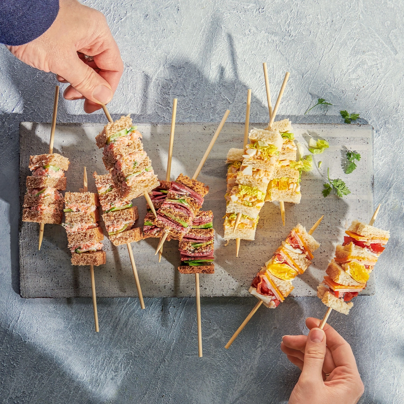 Köstliche Sandwich-Spieße auf einem grauen Brett, von Händen gehalten.