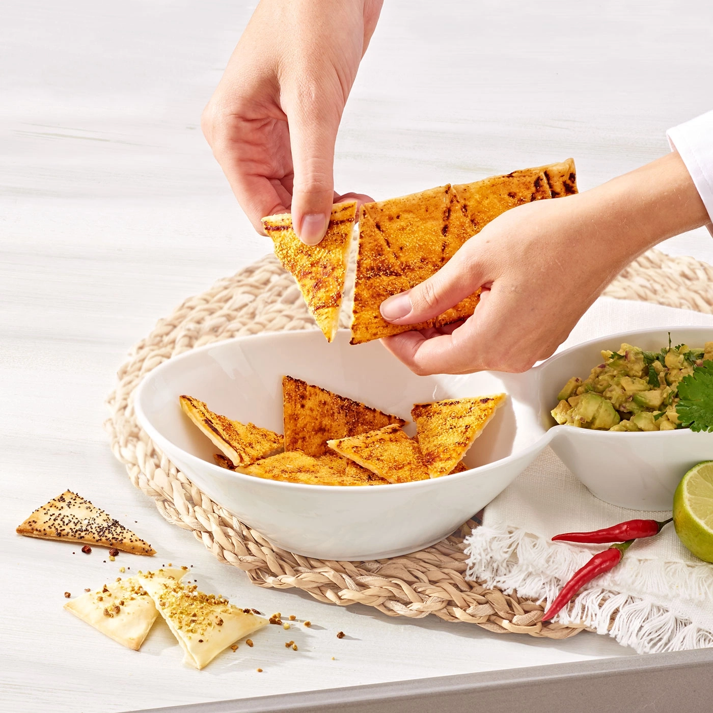 Hände halten Tortilla-Chips, Guacamole, Chili, Limette, Korbuntersetzer, weißer Tisch