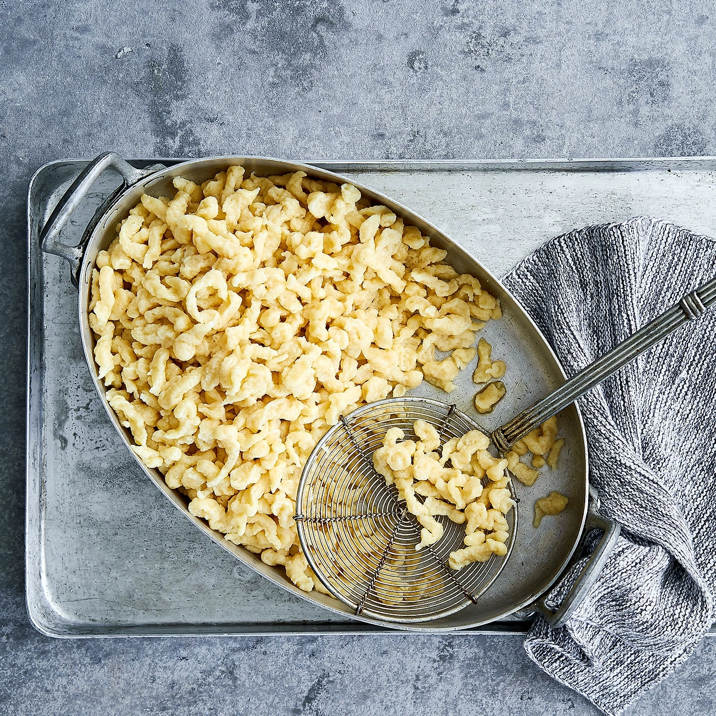 Spätzle in einer ovalen Schüssel mit einer Siebkelle und einem grauen Tuch auf einem Tablett