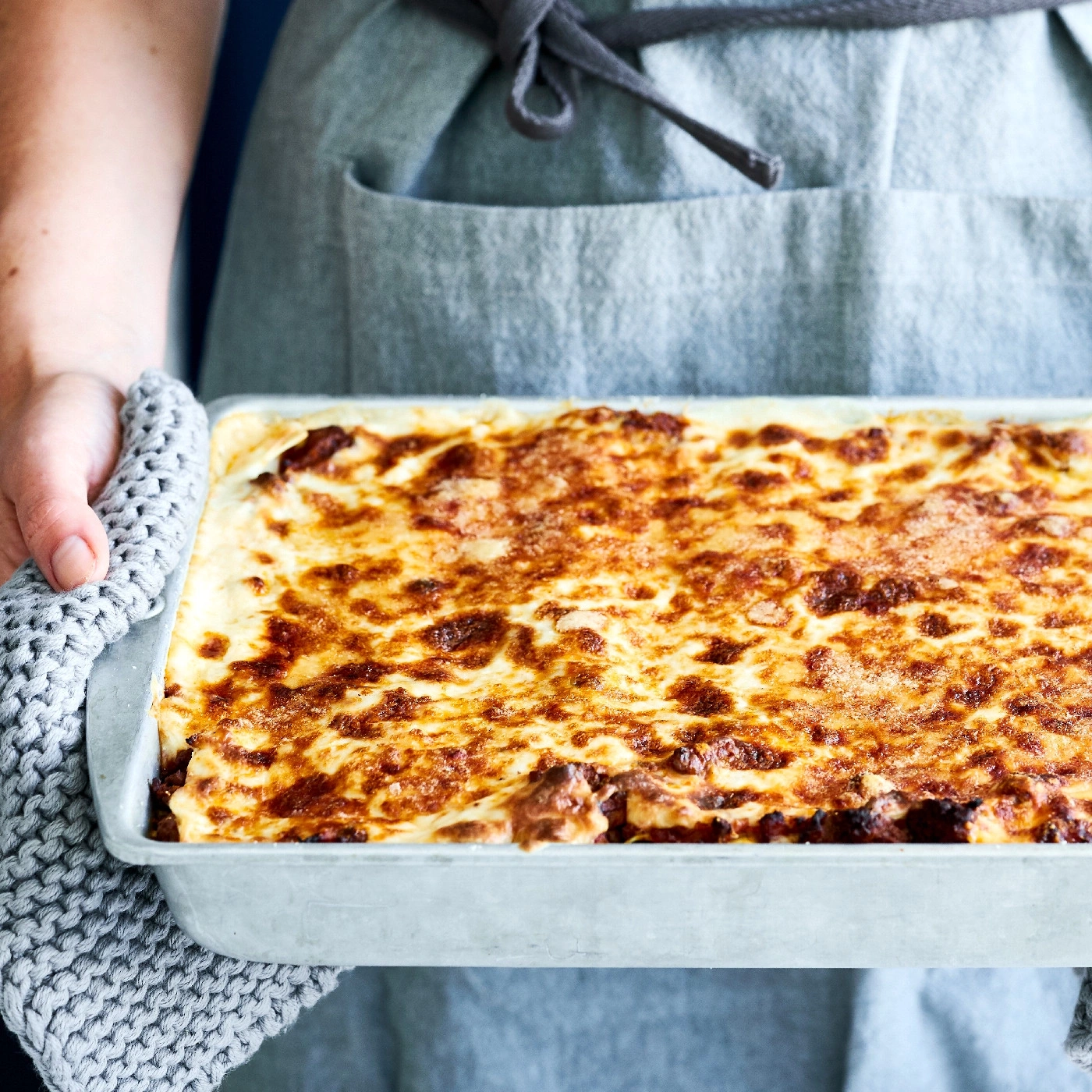 Lasagne in einer Backform, gehalten von einer Person mit einem grauen Topflappen.