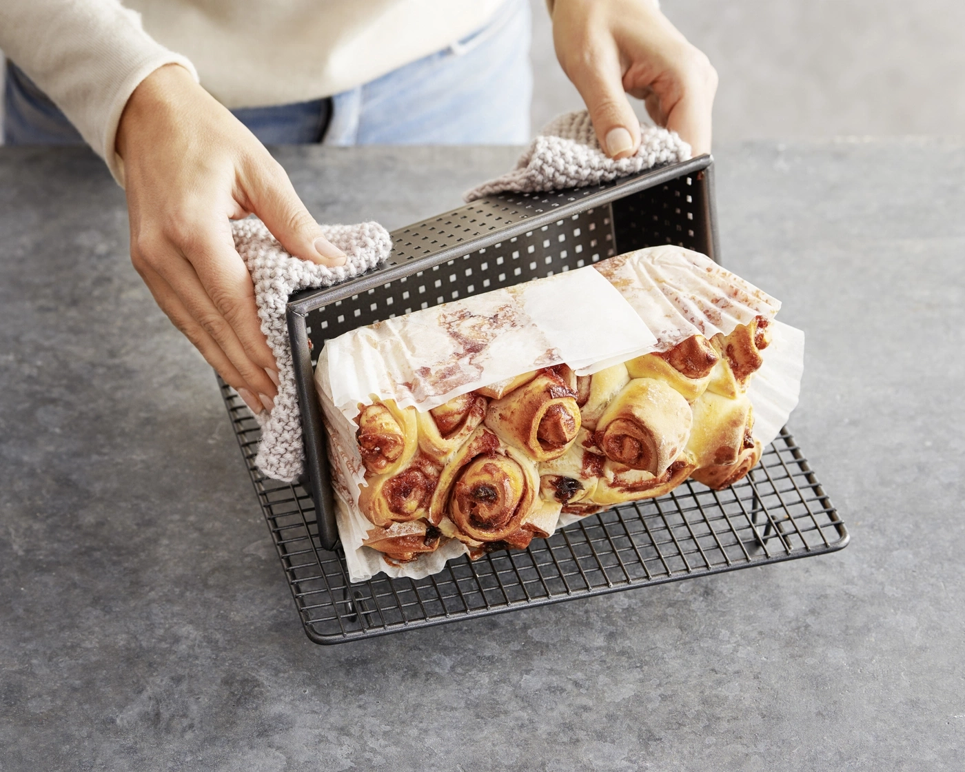Person hält Brot aus dem Ofen, Backform und Rost auf grauem Untergrund