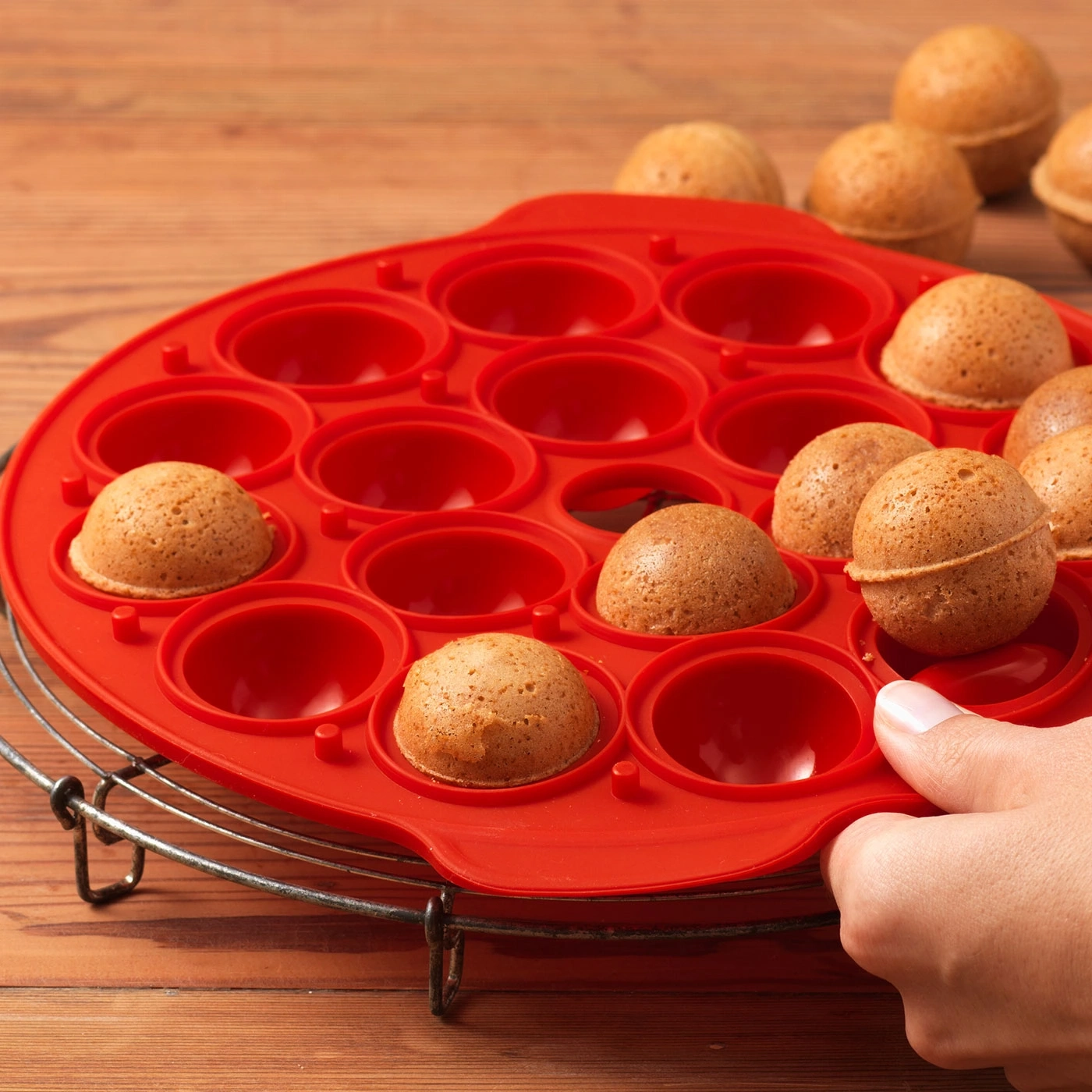 Rote Backform mit kleinen Kuchen, die von einer Hand gehalten werden, auf Holzuntergrund.