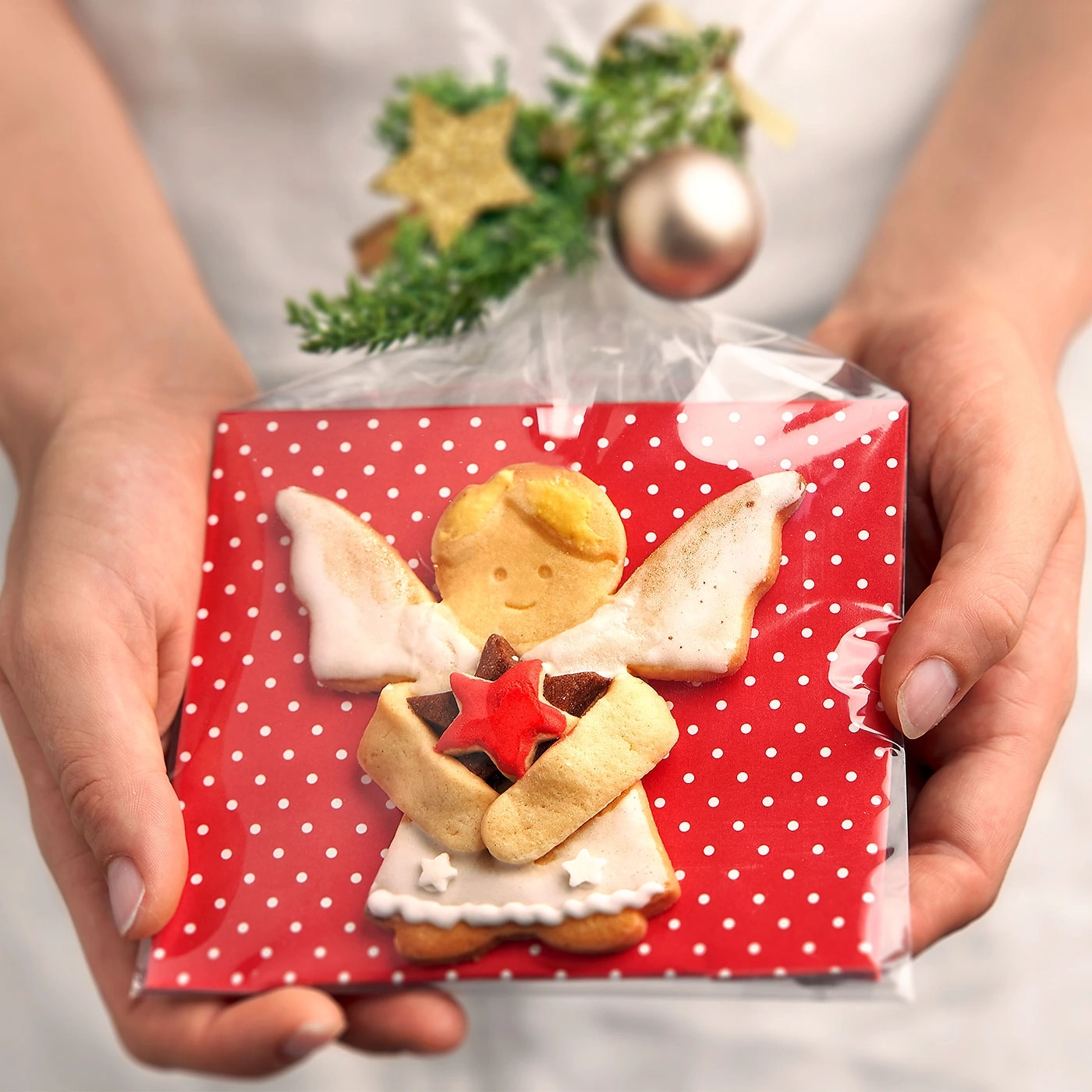 Personne tenant un cadeau avec un biscuit ange, papier rouge et pois blancs.