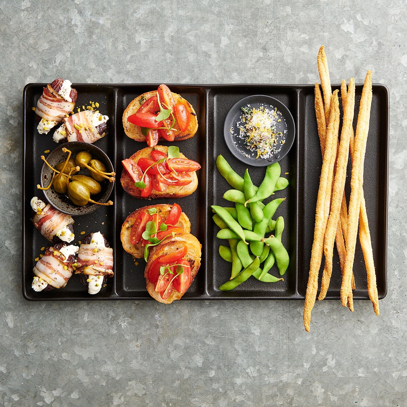 Tablett mit Snacks: Bruschetta, Edamame, Brotstangen, Kapern, Speck