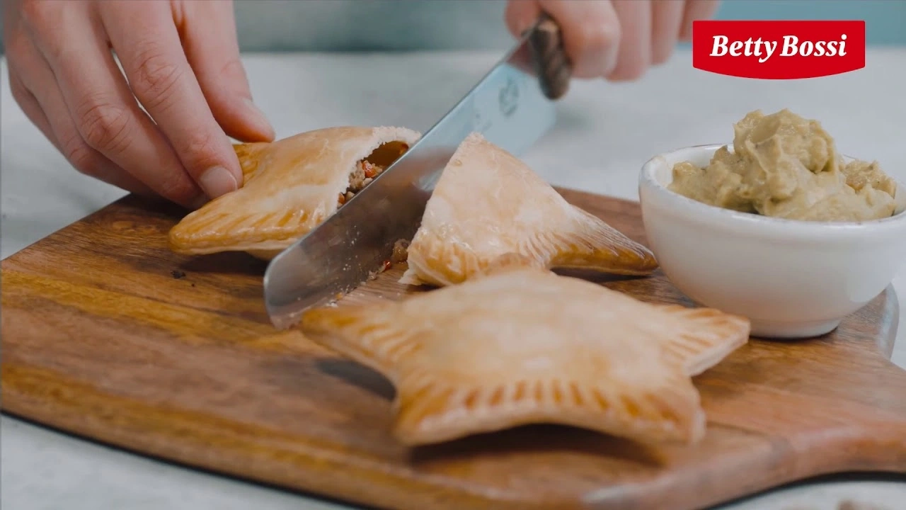 Personne coupant une pâtisserie sur une planche de bois avec une trempette dans un bol.