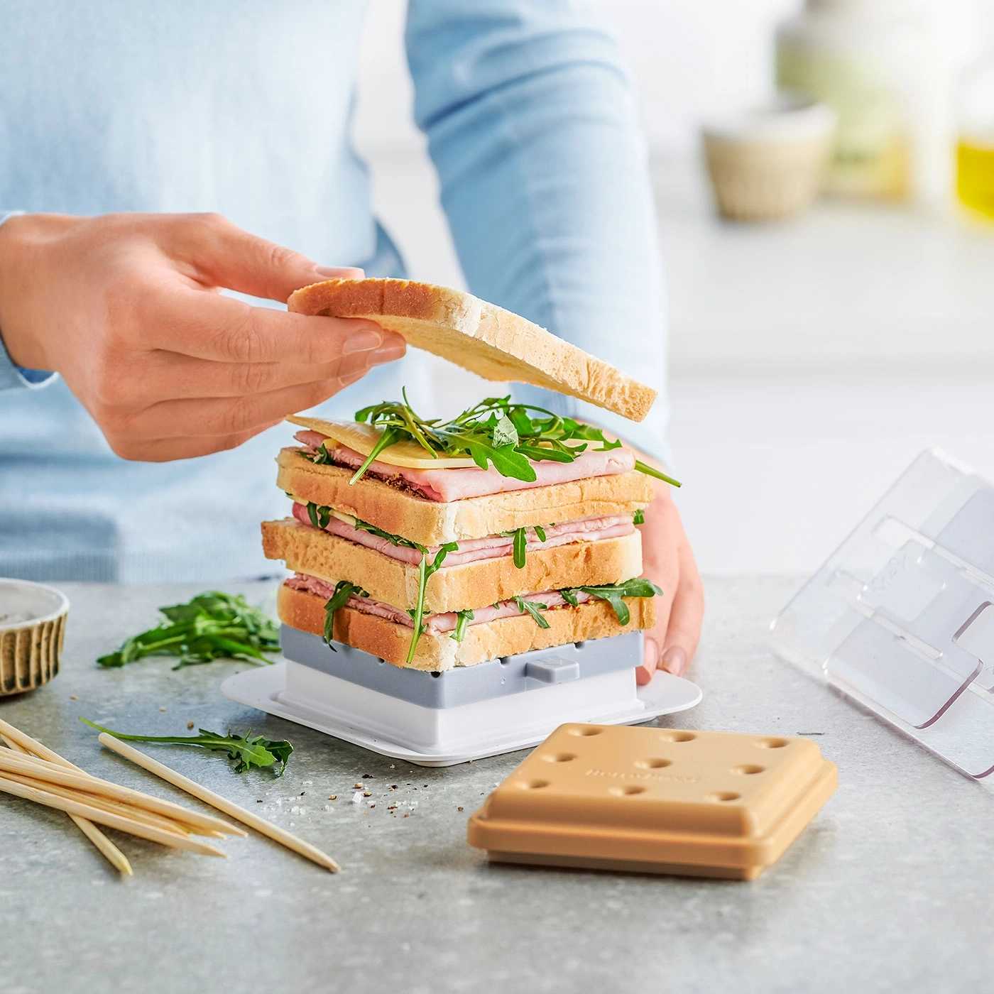 Person legt Sandwich-Scheibe auf einen Sandwich-Turm mit Käse und Rucola.
