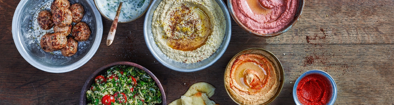 Verschiedene Gerichte auf einem Holztisch: Hummus, Salat, Fleischbällchen, Brot, Dips.
