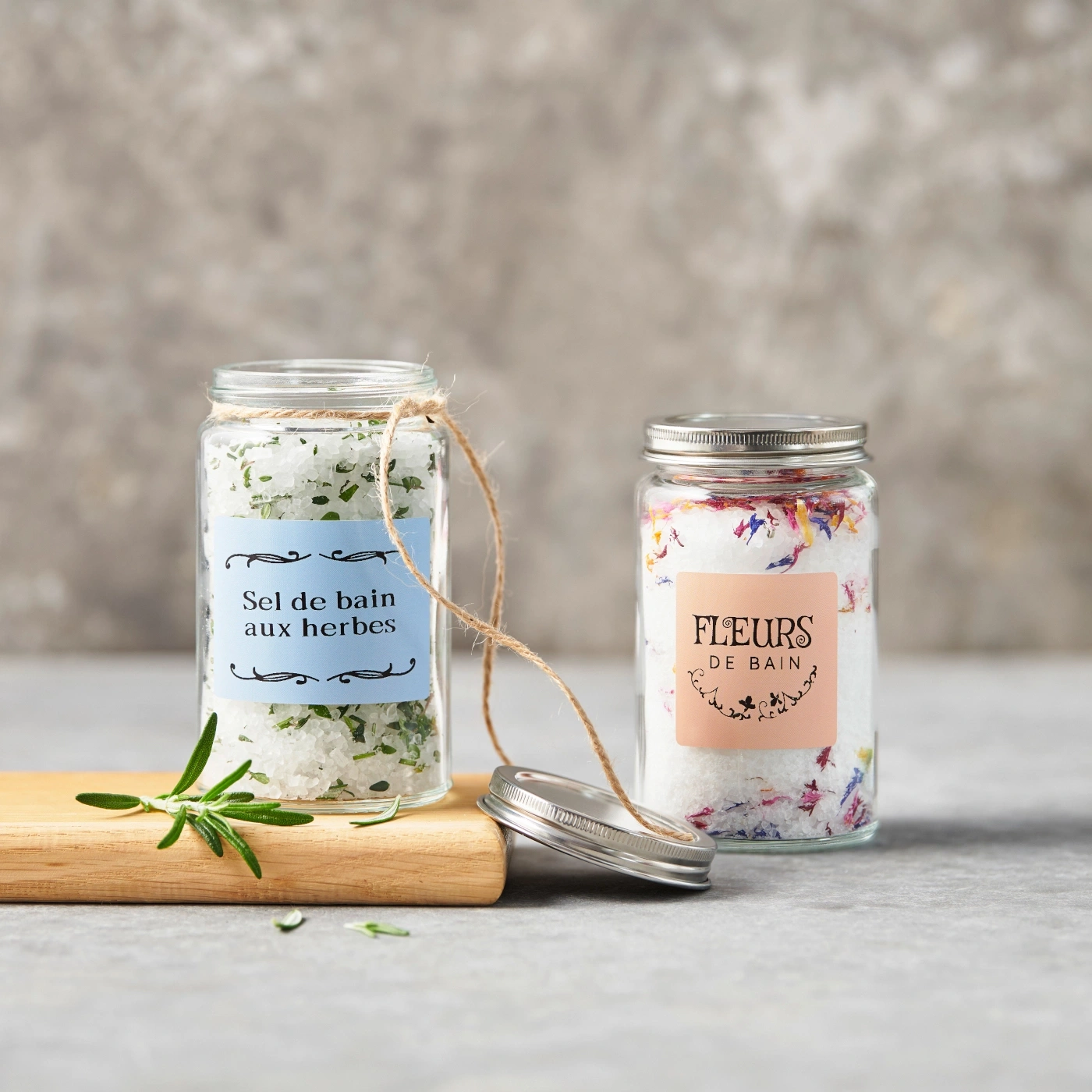 Deux pots de sels de bain, un aux herbes, un aux fleurs, sur une planche en bois.