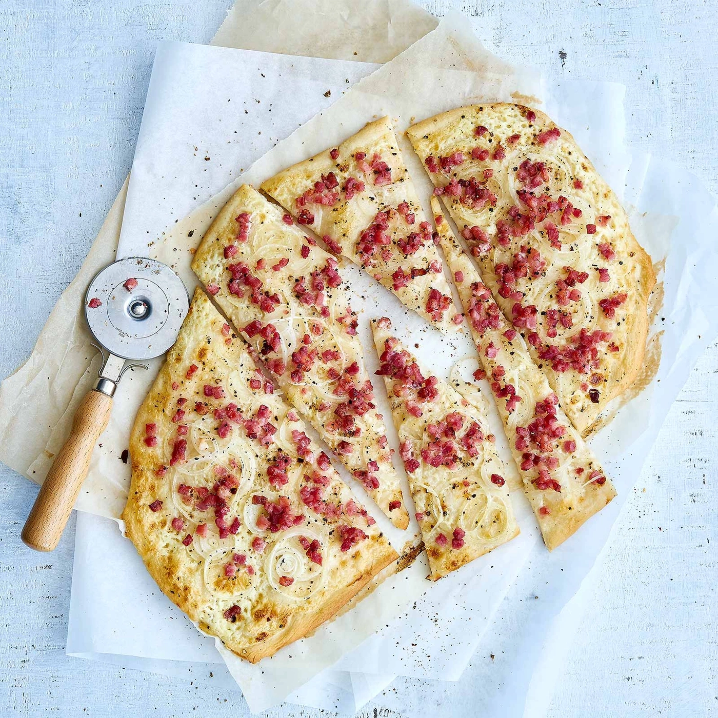 Flammkuchen, geschnitten, mit Zwiebeln und Speck auf Backpapier, Holzuntergrund