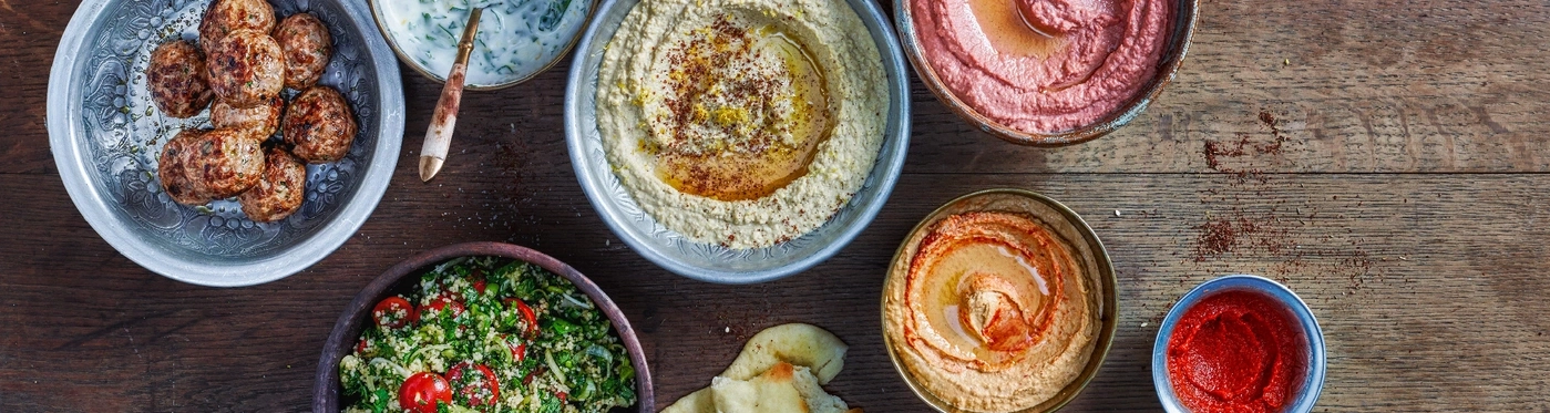 Verschiedene Gerichte auf einem Holztisch: Hummus, Salat, Fleischbällchen, Brot, Dips.