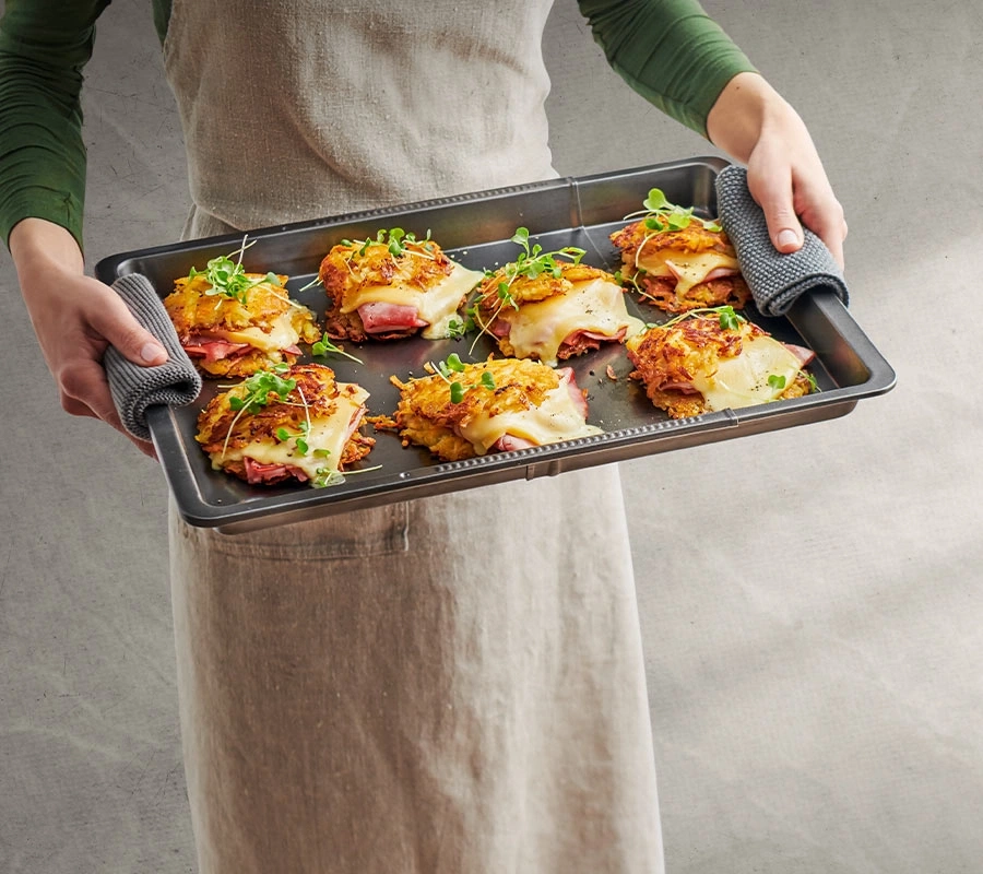 Person hält Backblech mit Essen. Käse, Schinken und Kartoffeln.