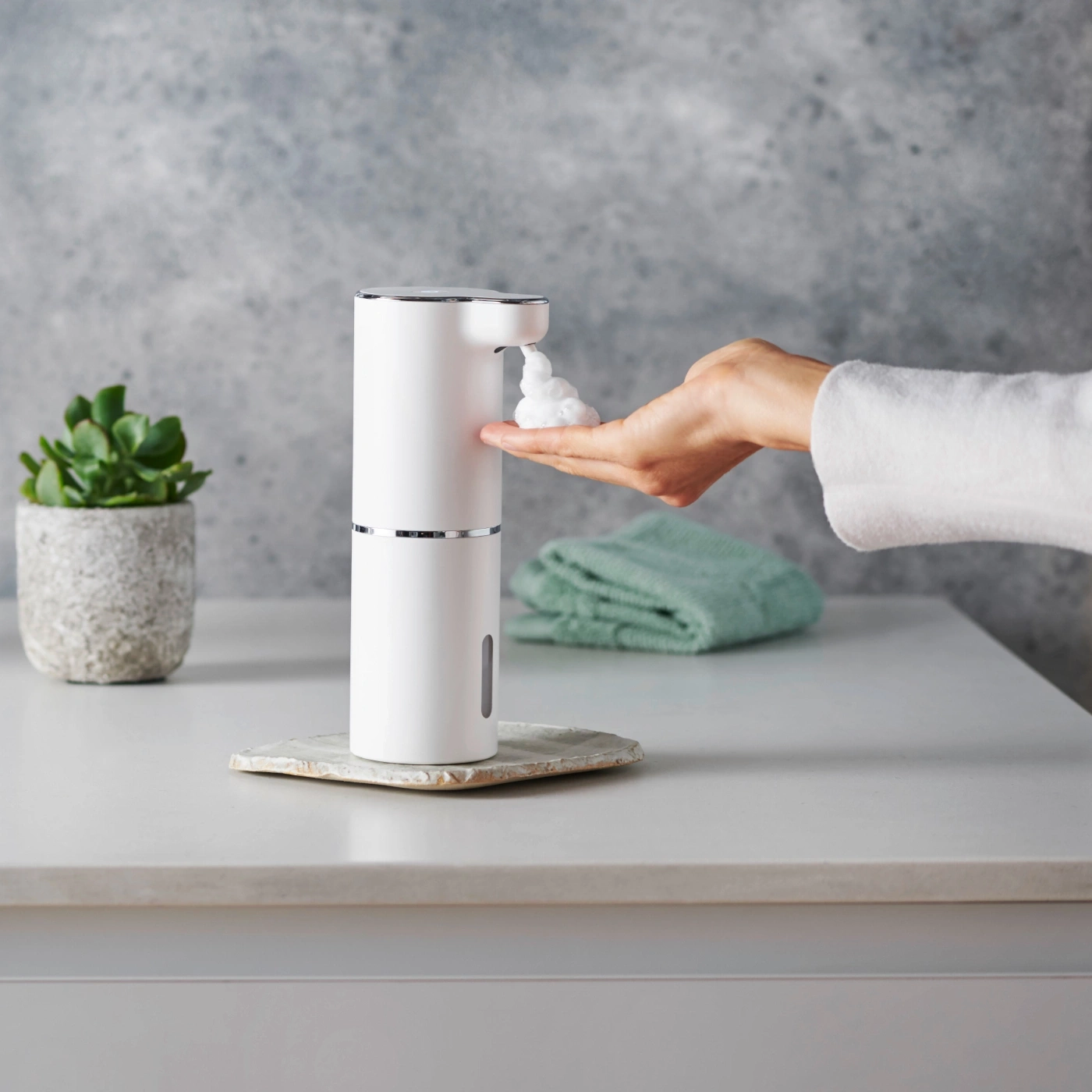 Distributeur de savon blanc, main avec mousse, plante, serviette, fond gris, salle de bain