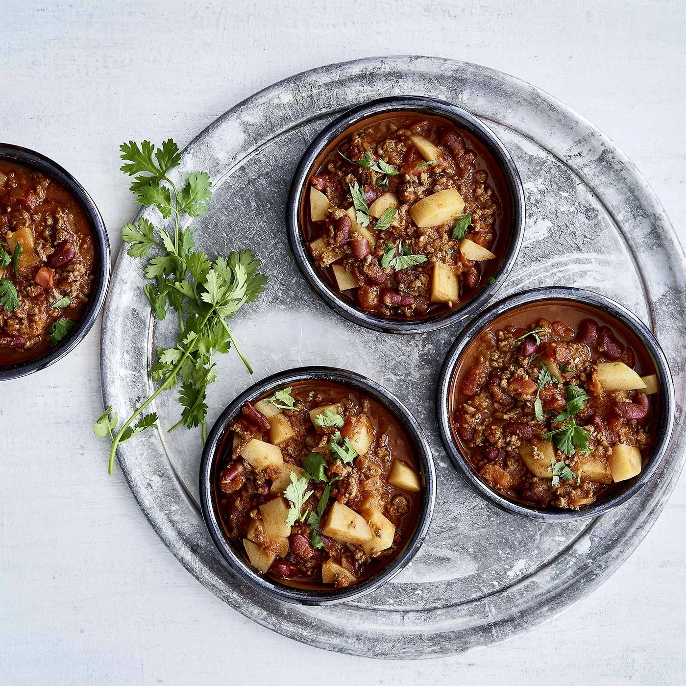 Chili-Gericht in Schalen auf einem Tablett mit Kräutern und Kartoffeln.