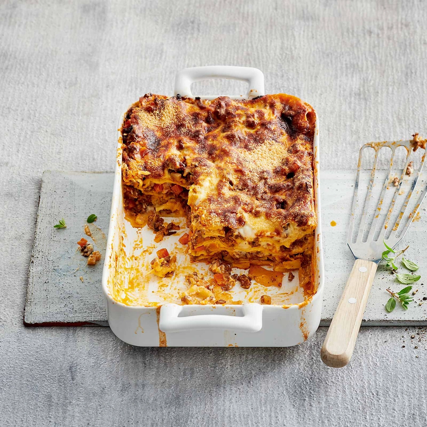 Lasagne in einer Auflaufform, serviert mit einem Spatel und Kräutern.