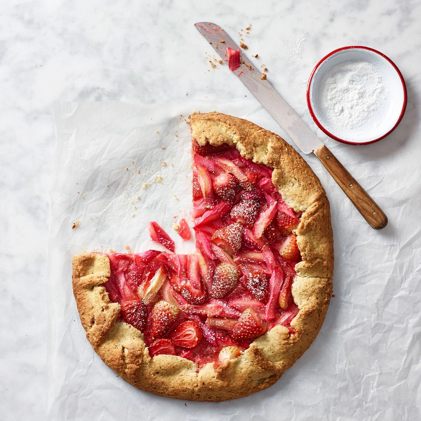 Erdbeer-Rhabarber-Tarte mit Puderzucker, Messer und Schüssel auf Papier