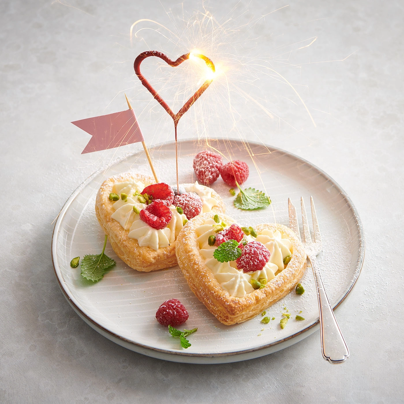 Herzförmige Gebäckstücke mit Himbeeren, Sahne und einem Herz-Sparkler auf einem Teller.