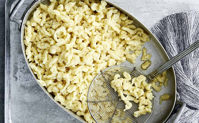 Spätzle in einer Schüssel mit einem Sieblöffel und einem grauen Tuch.