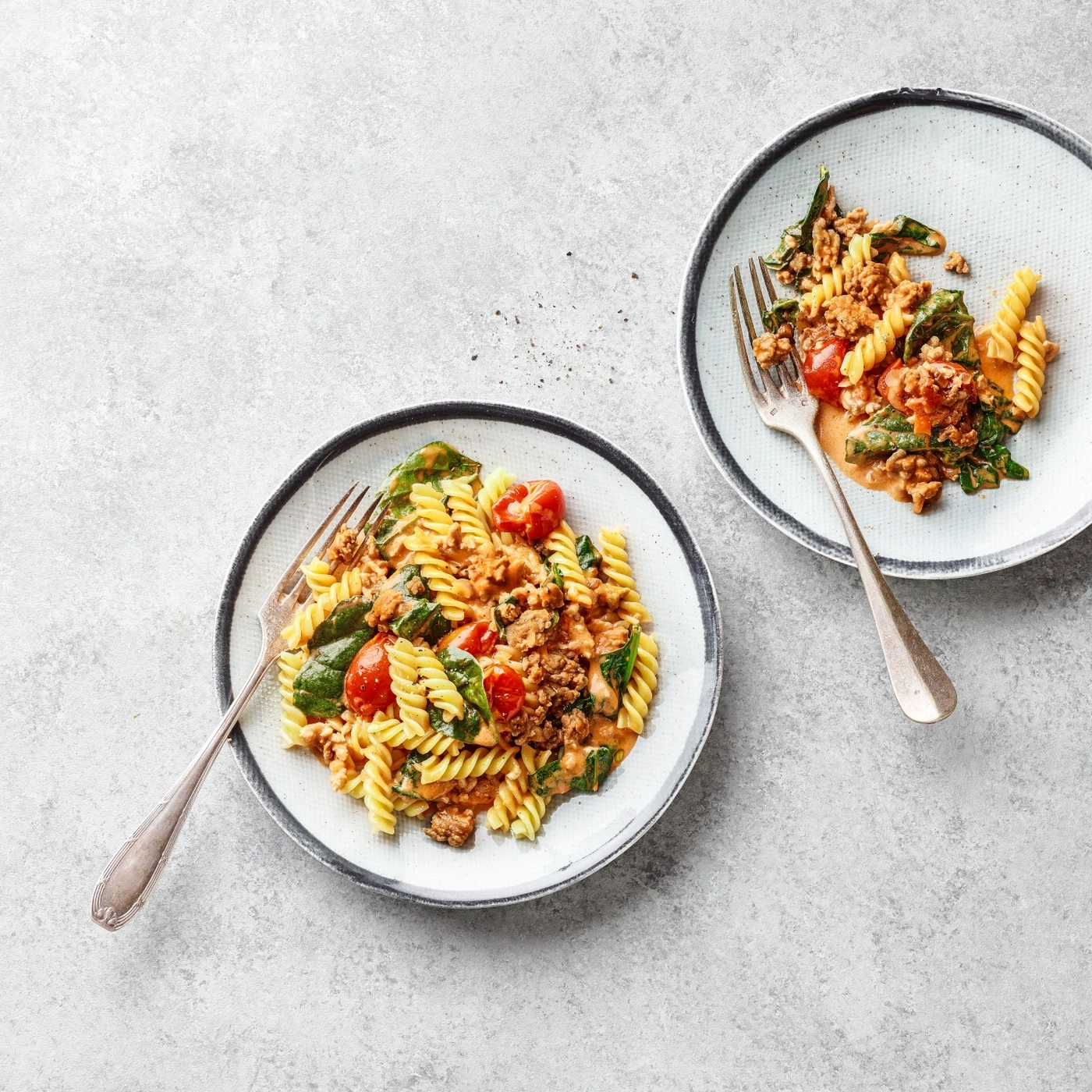 Deux assiettes de pâtes avec tomates et viande sur fond gris.
