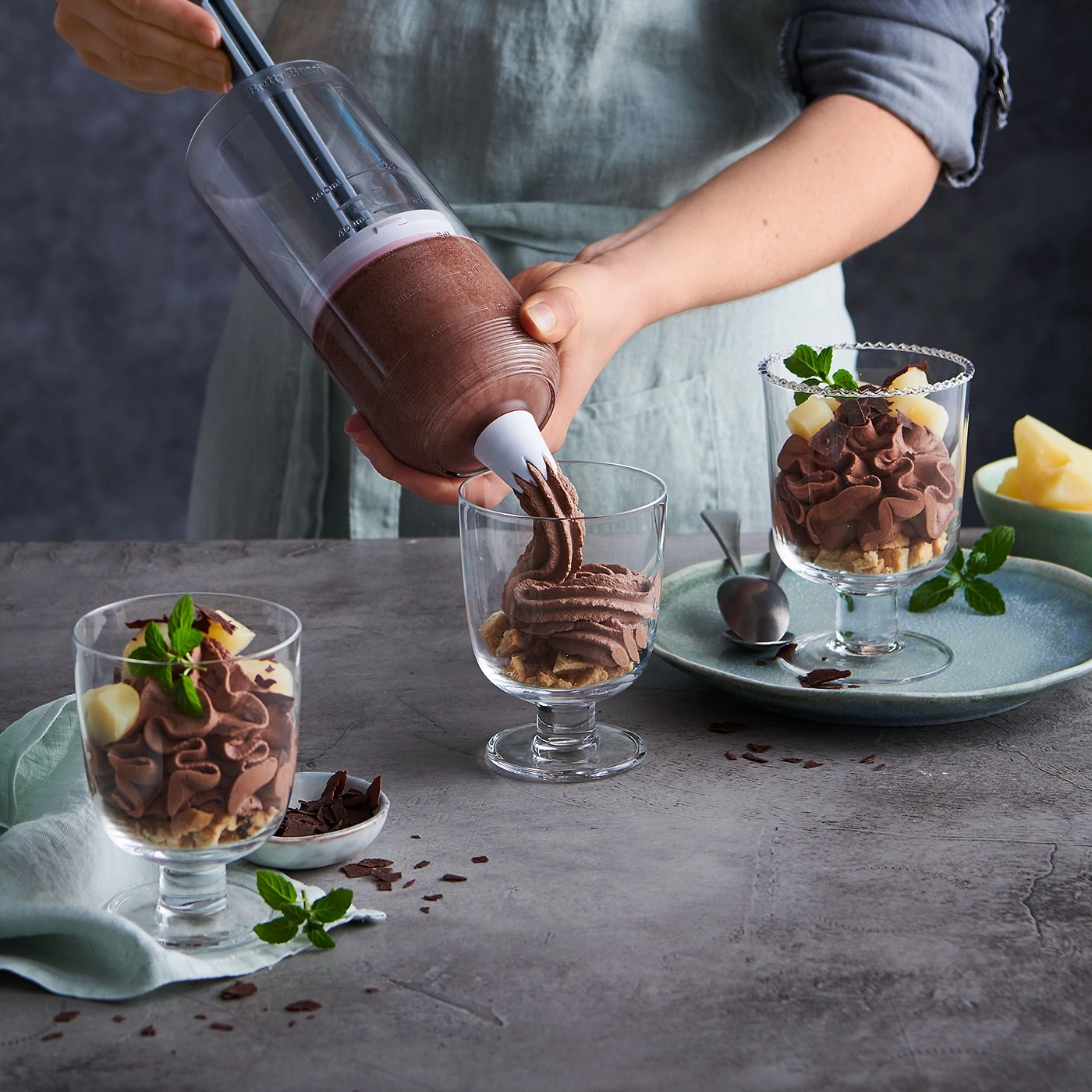 Personne versant mousse au chocolat dans des verres, garnie de menthe et de fruits.