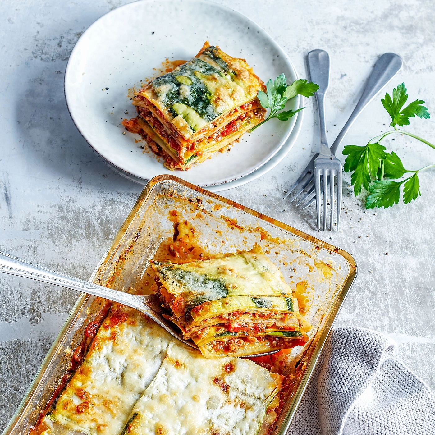Lasagne auf einem Teller und in einer Auflaufform mit Kräutern und Gabeln