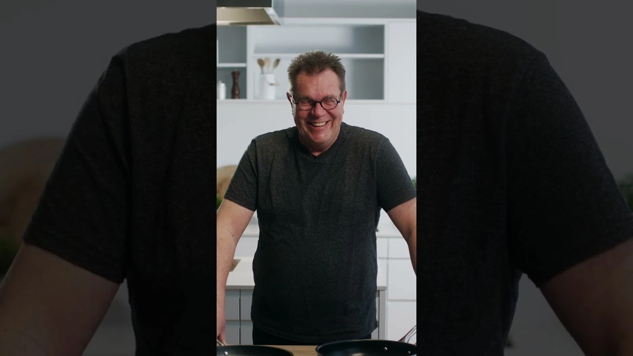 Ein Mann mit Brille und grauem T-Shirt lächelt in einer Küche mit Pfannen.