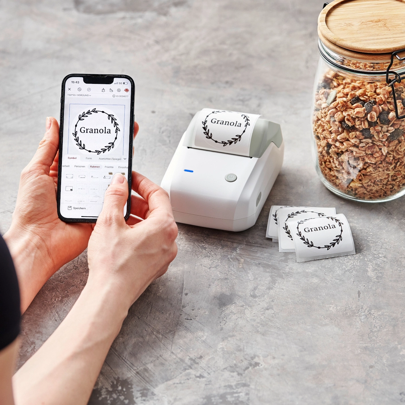 Person hält Smartphone, Etikettendrucker, Granola-Etiketten, Glas mit Granola, grauer Hintergrund.