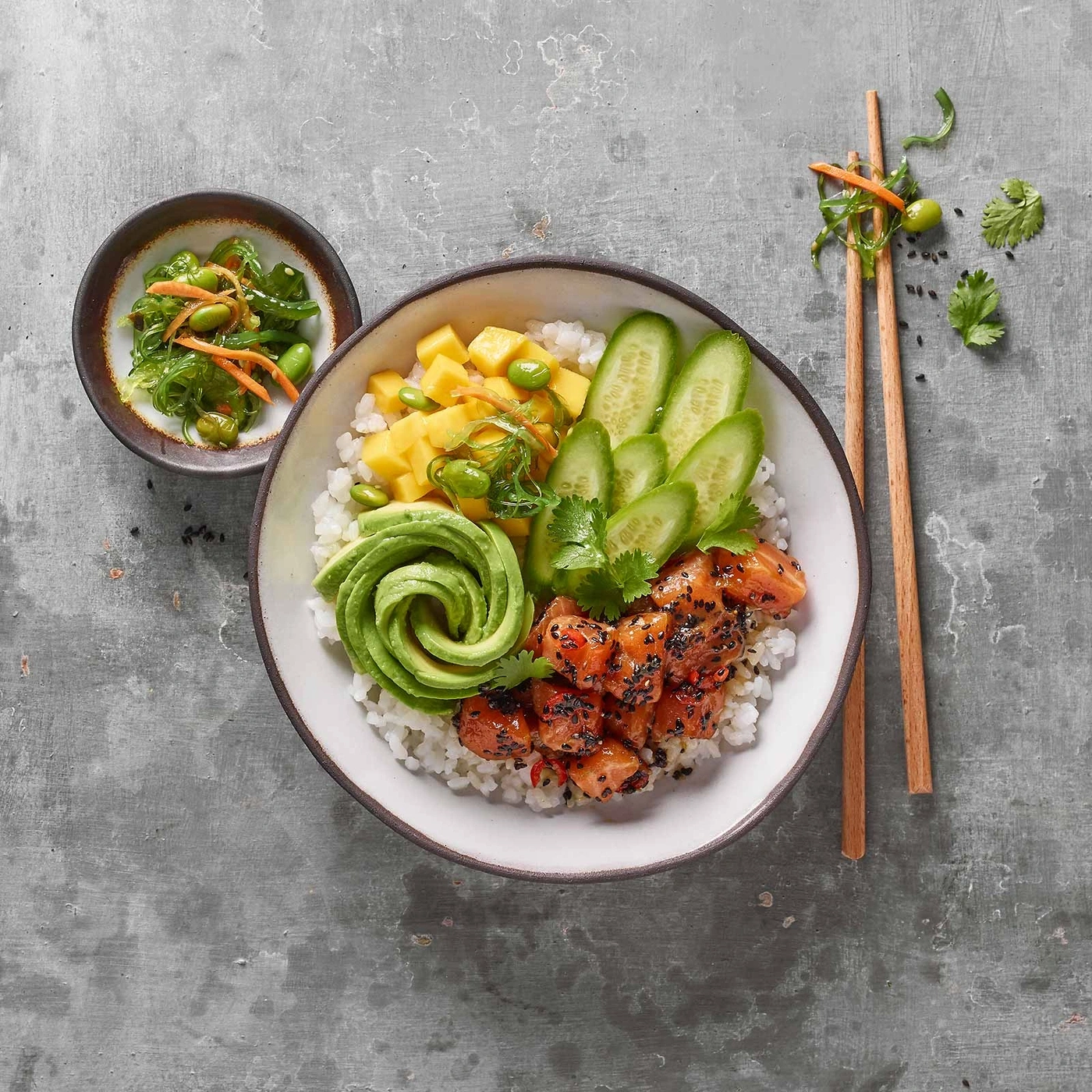 Poke Bowl mit Lachs, Avocado, Mango, Gurke und Reis, serviert mit Stäbchen.