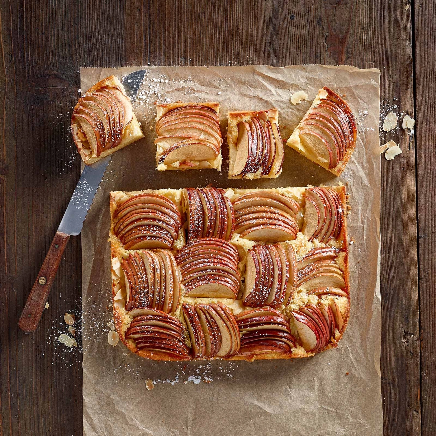 Apfelkuchen auf Backpapier mit Messer und Holzuntergrund, Ansicht von oben