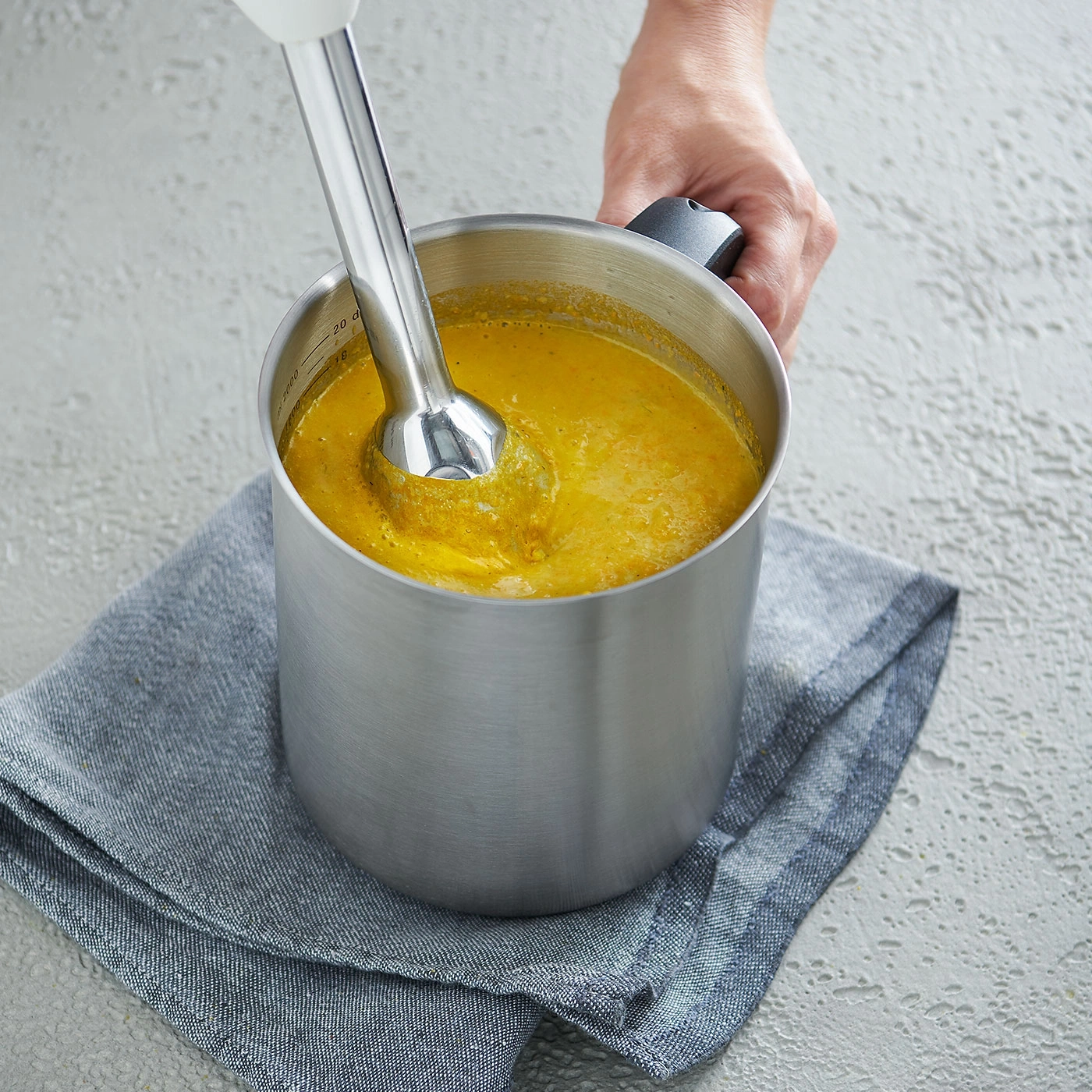 Person püriert Suppe mit einem Stabmixer in einem Topf auf einem grauen Tuch.