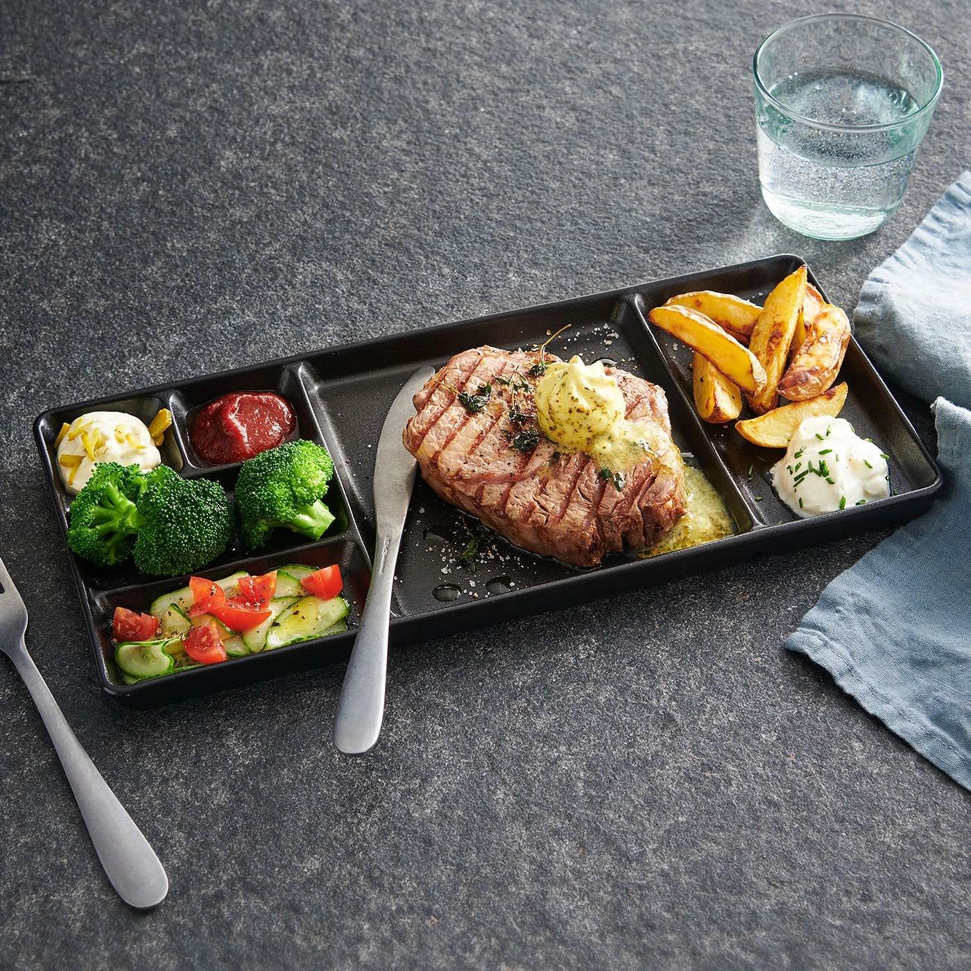 Steak avec accompagnements sur une assiette noire, verre d'eau et couverts.