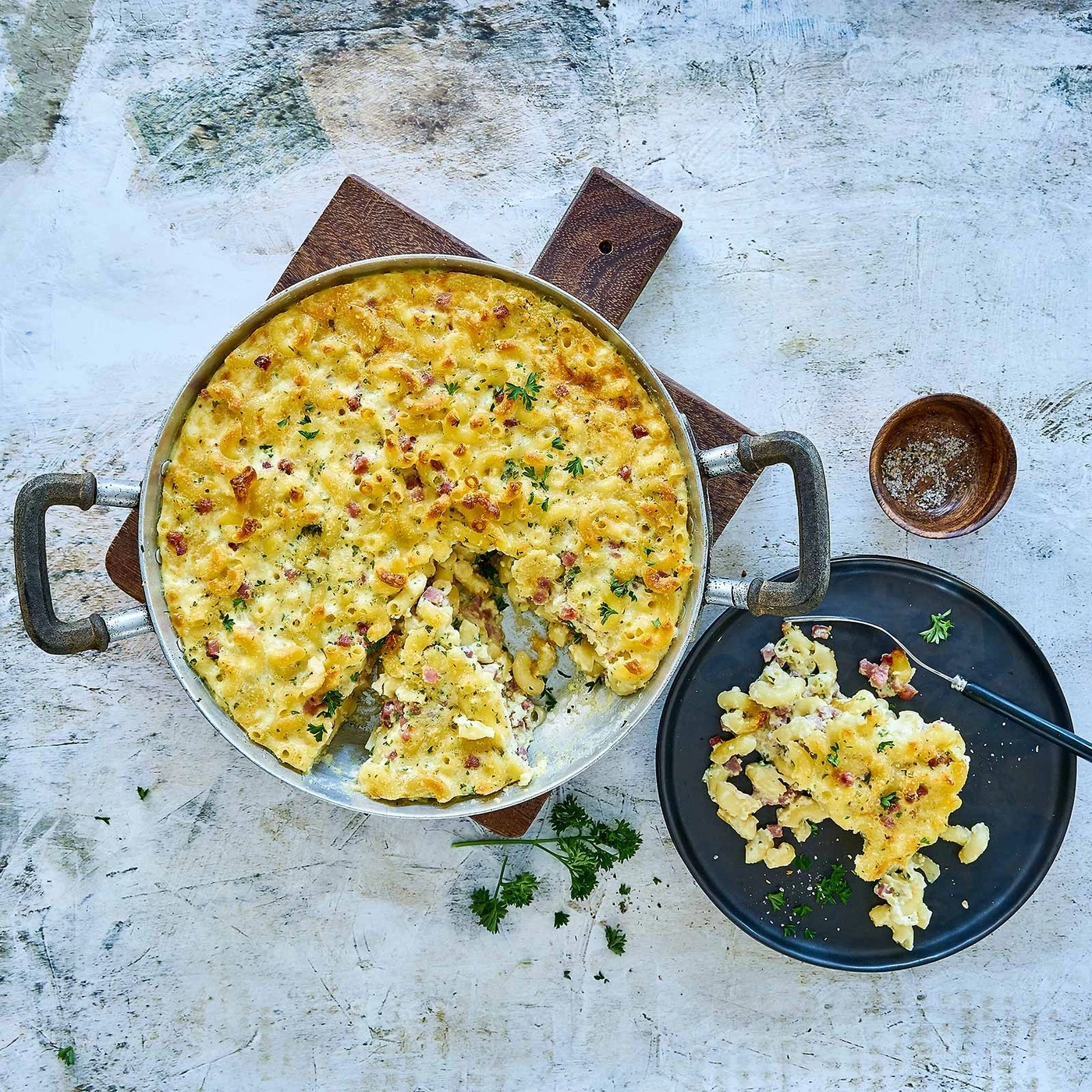 Käse-Makkaroni-Auflauf in Pfanne und auf Teller, mit Kräutern und Speck