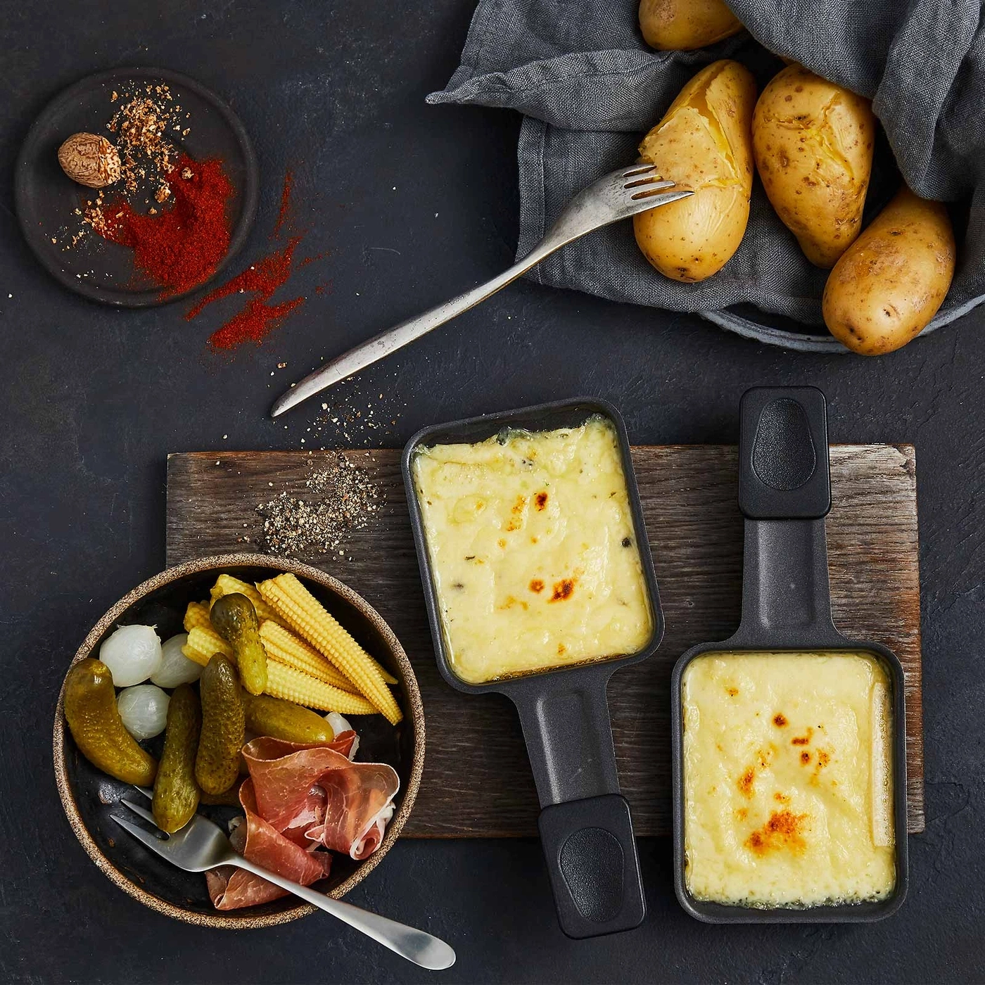 Raclette-Gericht mit Kartoffeln, Käse, Gemüse und Gewürzen auf dunklem Hintergrund.