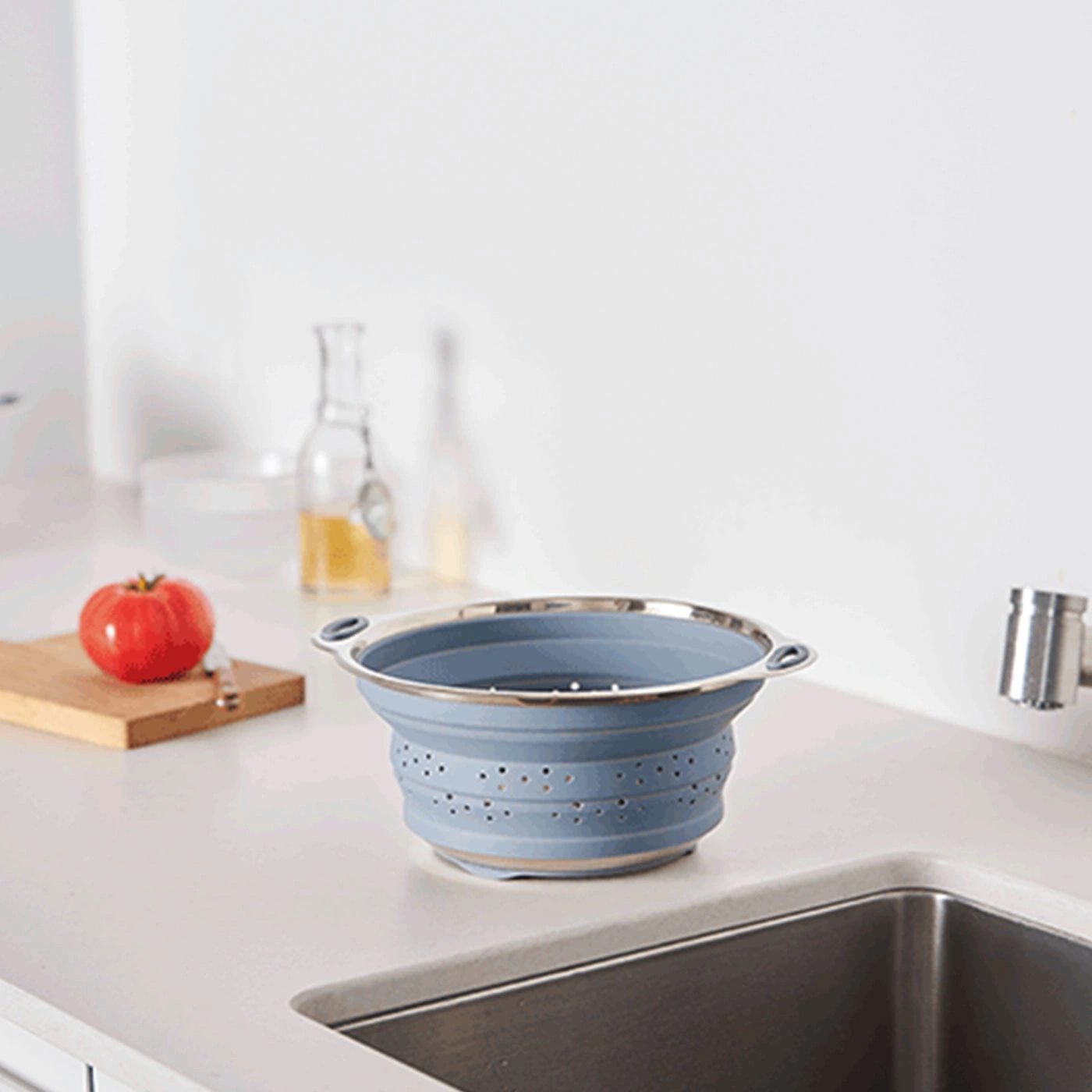 Passoire de cuisine bleue pliable sur un comptoir blanc avec une tomate.