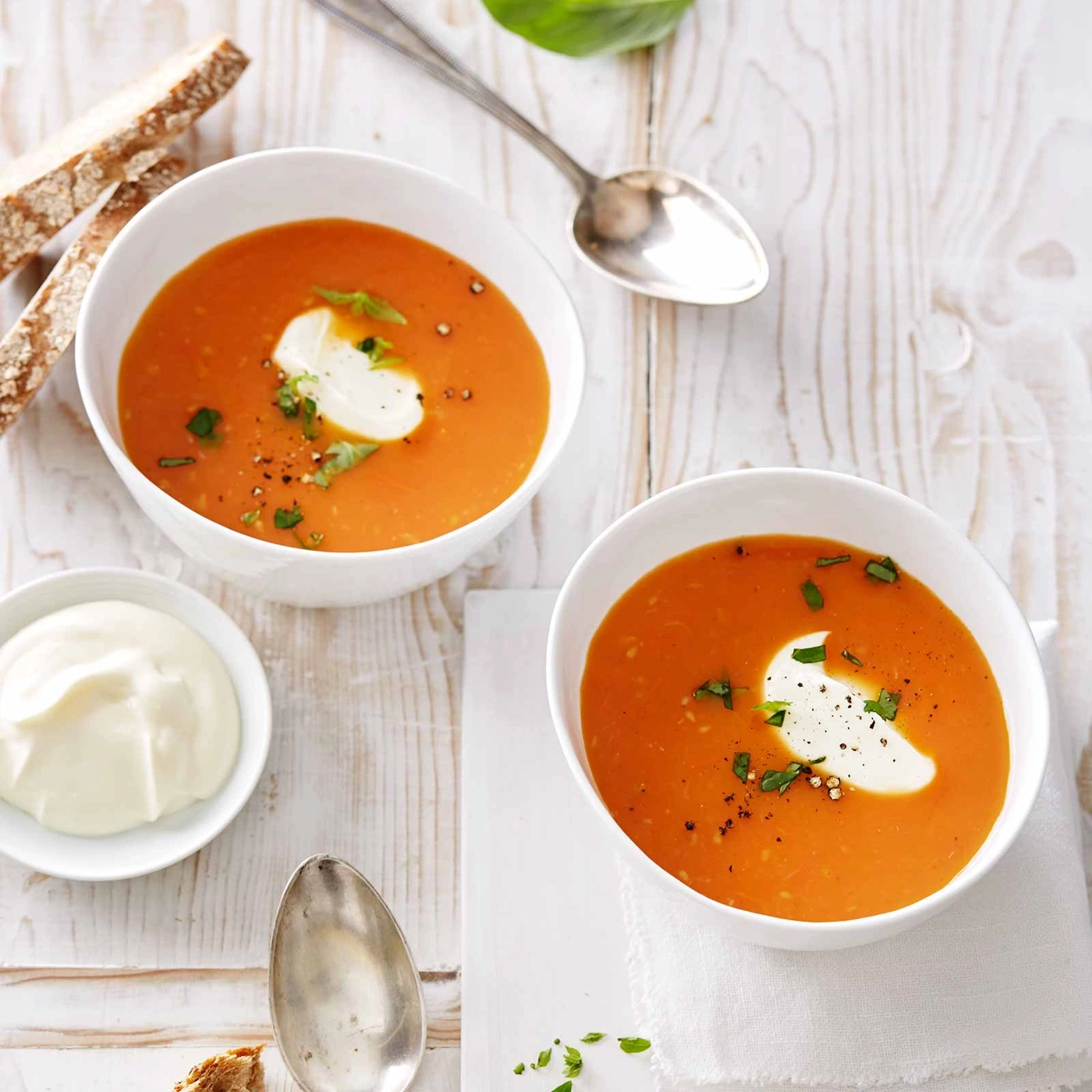 Zwei Schüsseln Tomatensuppe mit Sahne, Brot und Löffel auf Holz