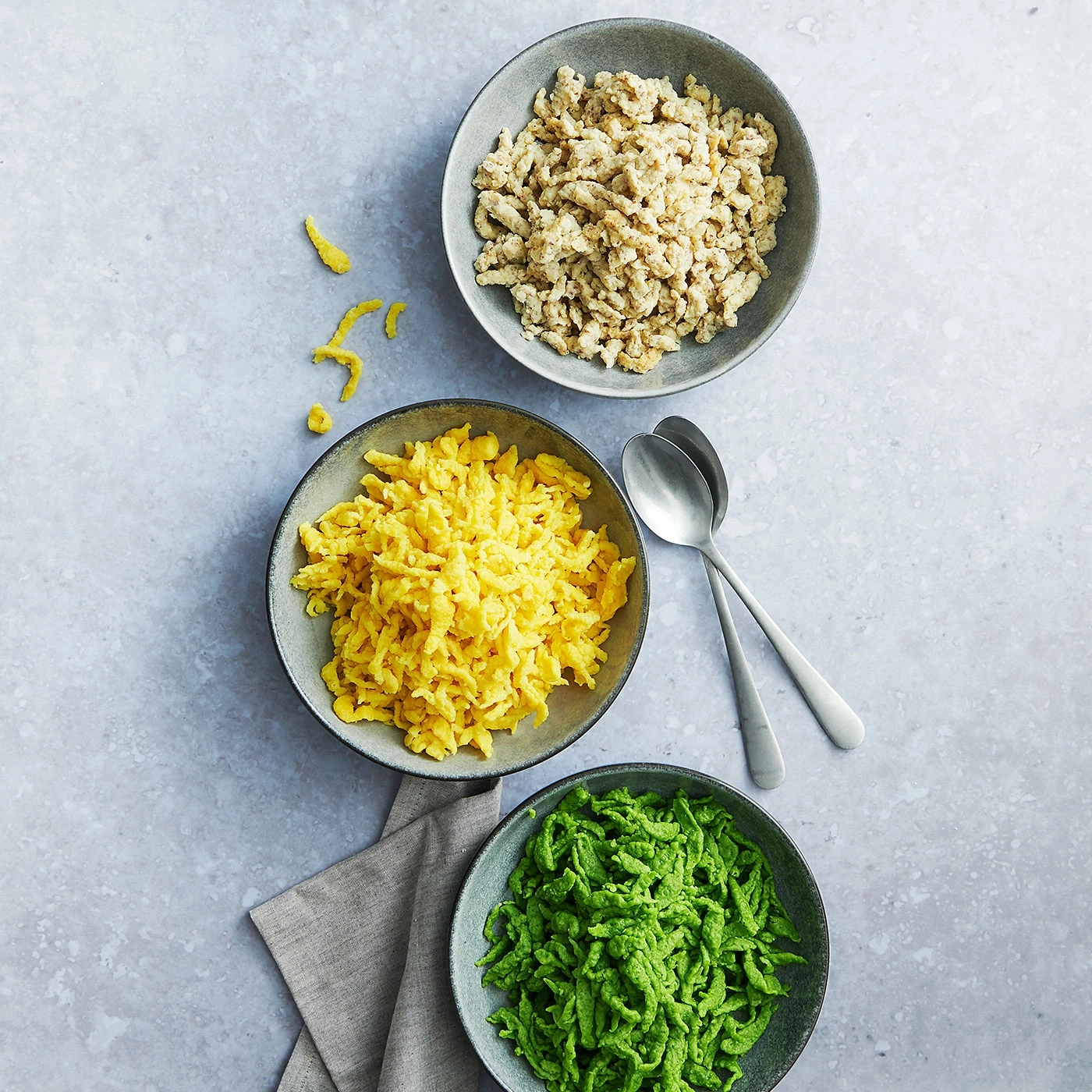 Drei Schüsseln Spätzle in verschiedenen Farben, mit Löffeln und einem Tuch.