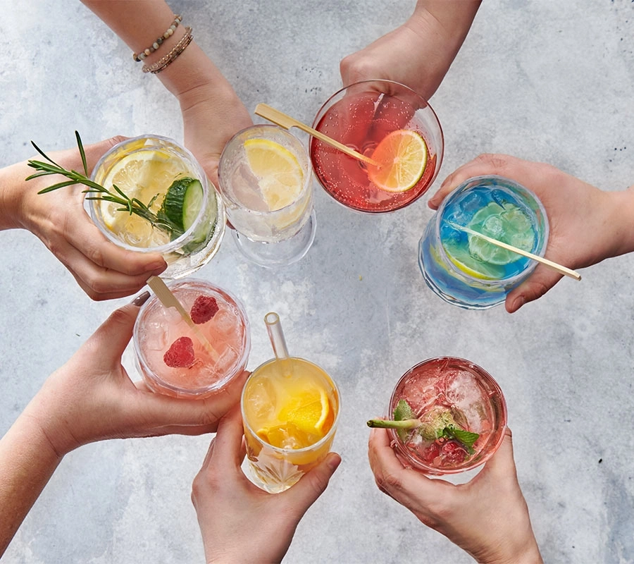 Hände halten Cocktails mit Früchten und Kräutern auf grauem Hintergrund.