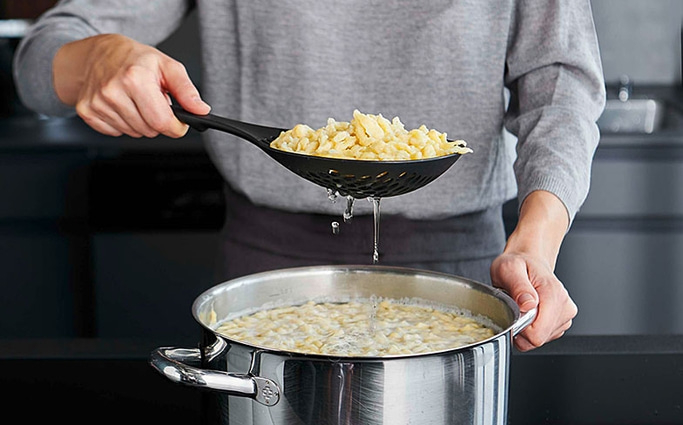Person kocht Spätzle, die aus dem Topf mit einem Schaumlöffel genommen werden.