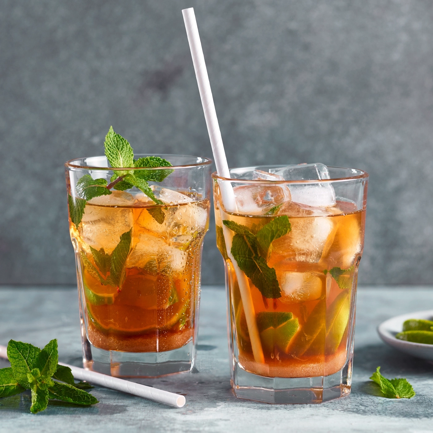 Zwei Gläser Eistee mit Minze, Limette und Eiswürfeln, mit Strohhalmen.