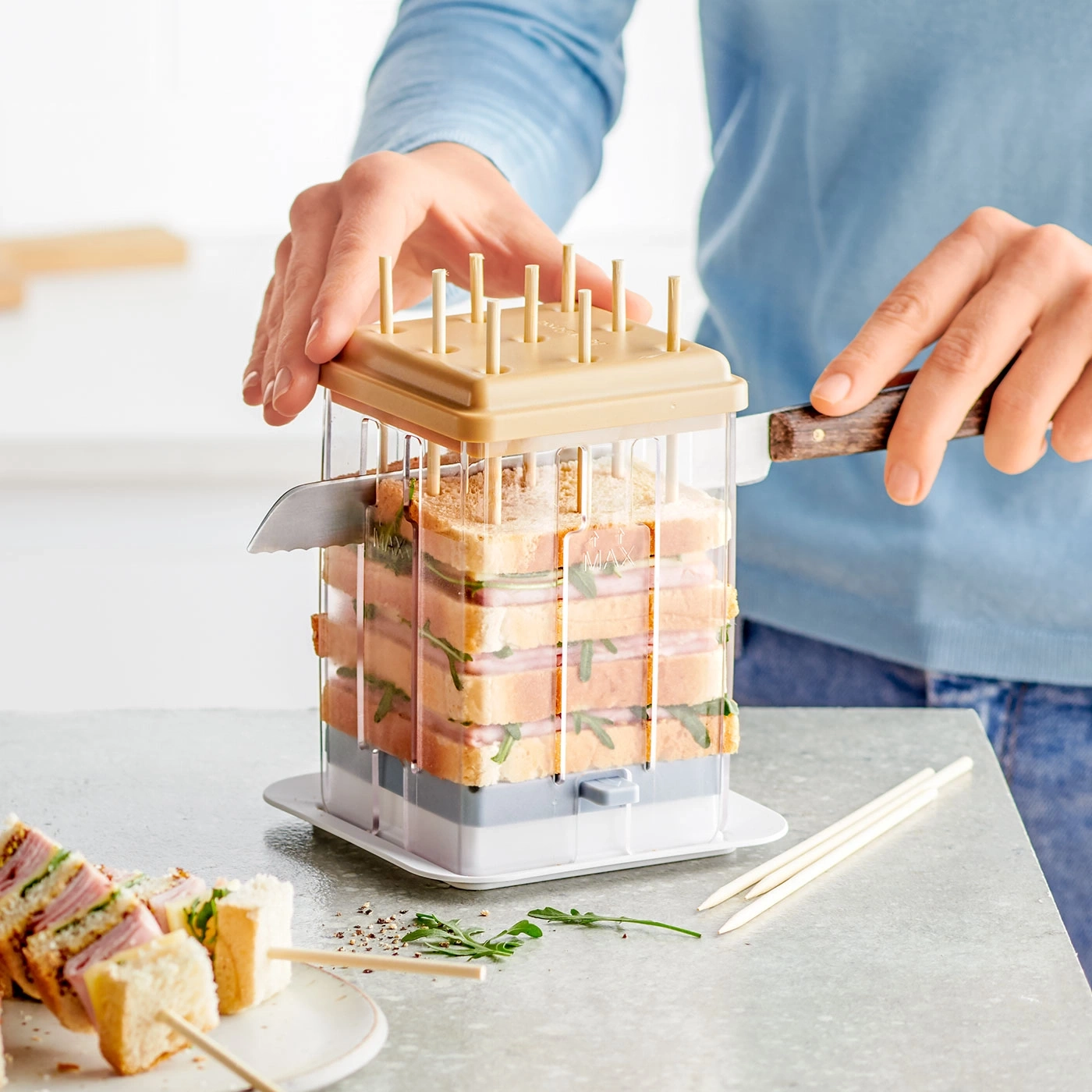 Person schneidet Sandwich mit Messer in Sandwichschneider, Sandwichspieße liegen daneben.