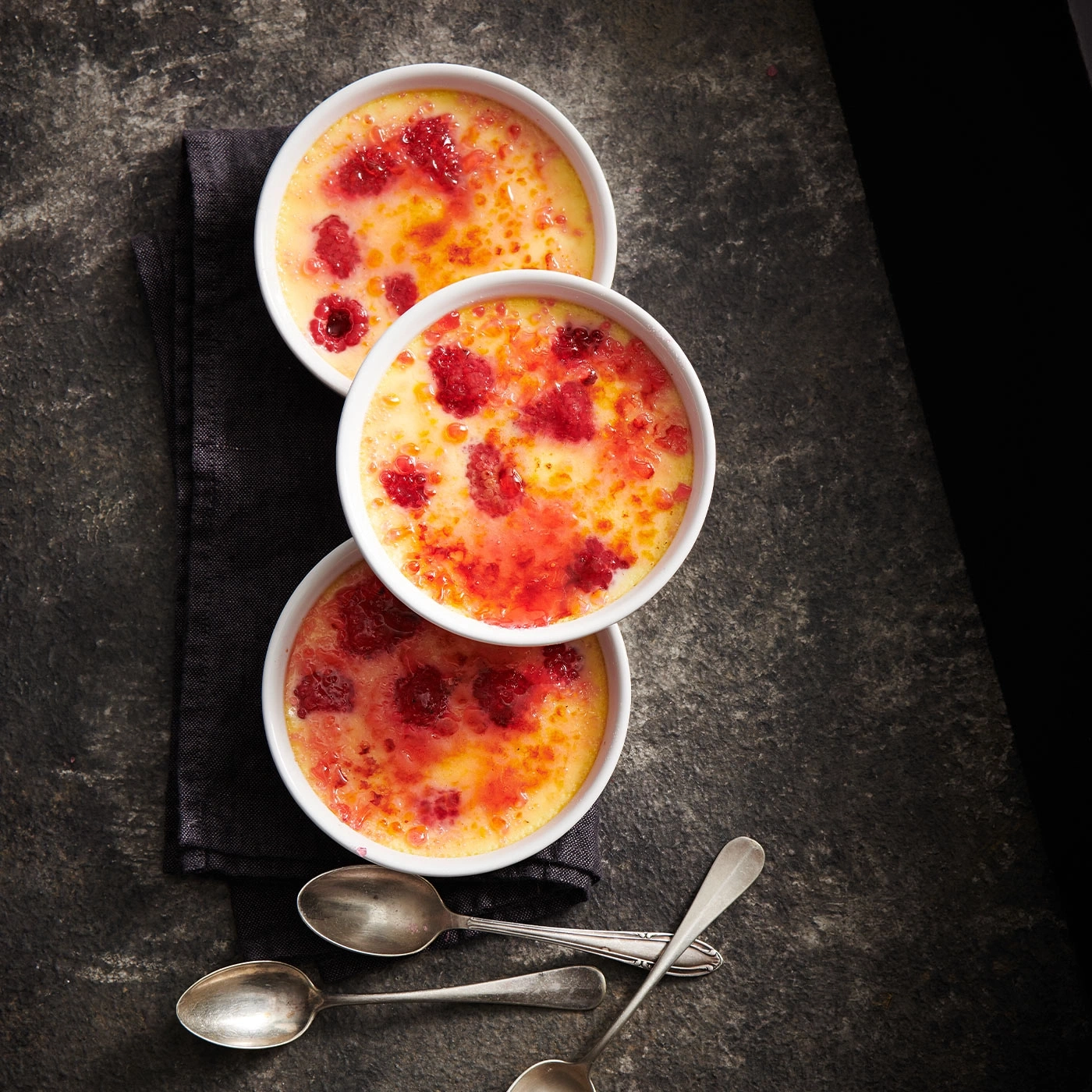 Drei Schalen Crème brûlée mit Himbeeren auf schwarzem Tuch und Löffeln.