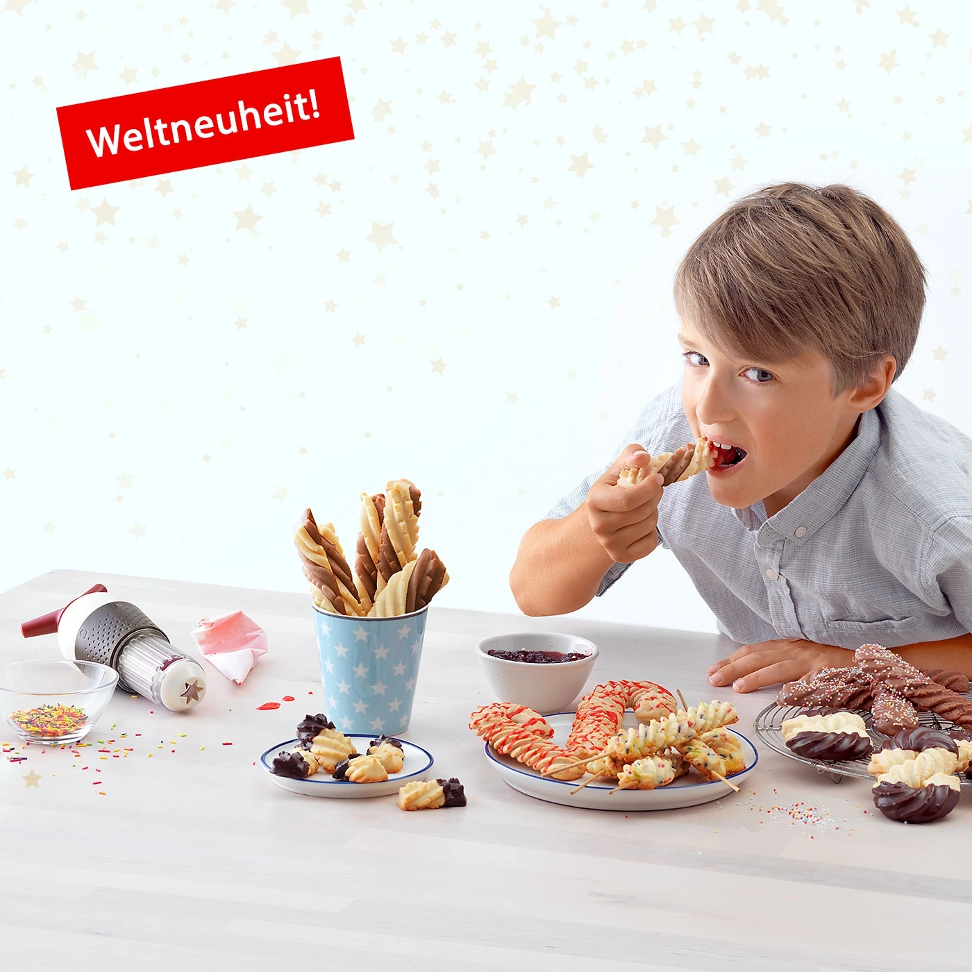 Junge isst Kekse. Auf dem Tisch sind Kekse, Streusel, eine Maschine und Marmelade.