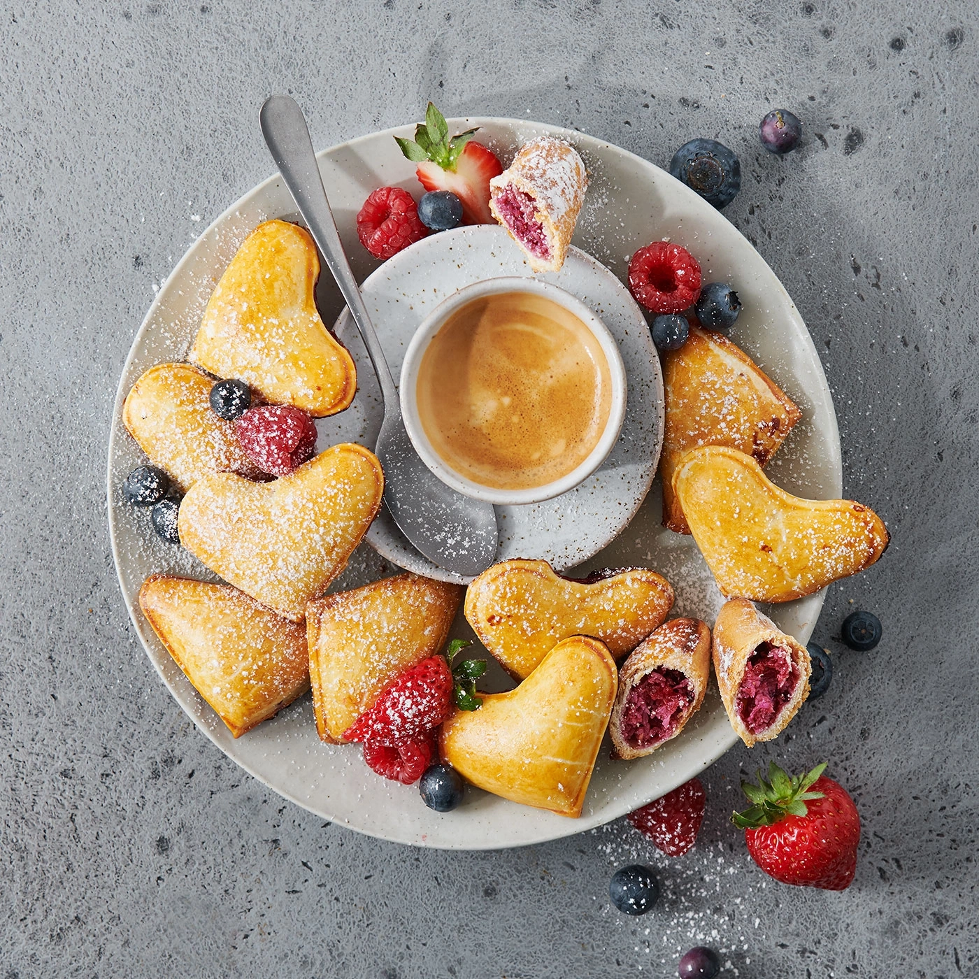 Herzförmiges Gebäck mit Beeren und Kaffee auf einem Teller.