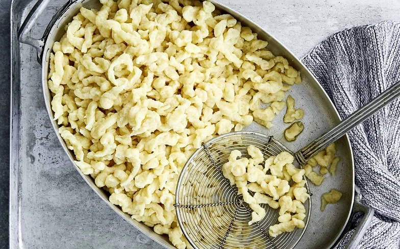 Spätzle in einer Schüssel mit einem Sieblöffel und einem grauen Tuch.