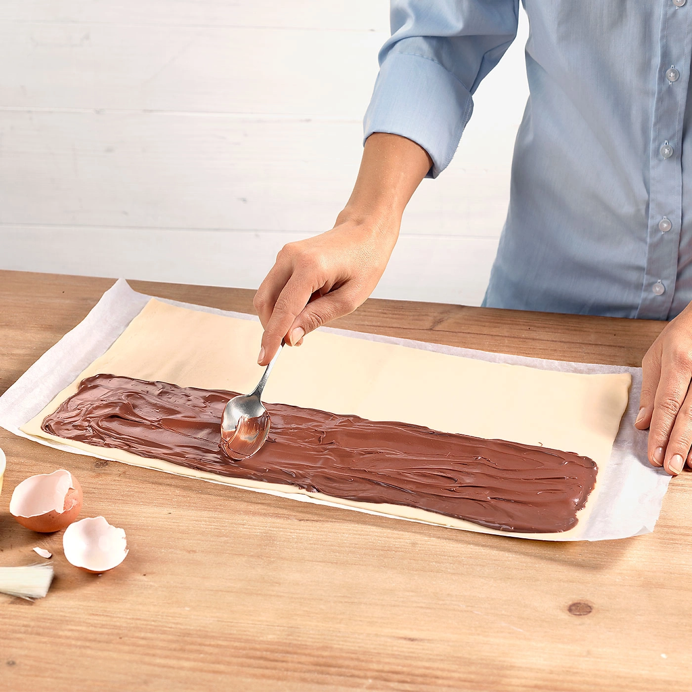 Person verteilt Schokoladencreme auf einem Teigblatt mit einem Löffel auf einem Holztisch.