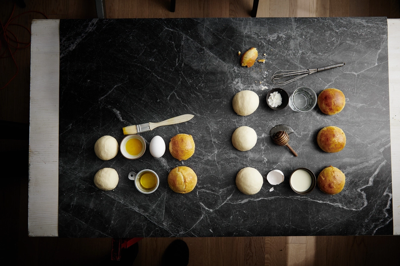 Backzutaten auf schwarzem Marmortisch: Teig, Eier, Öl, Milch, Honig, Wasser, Mehl, Brötchen.