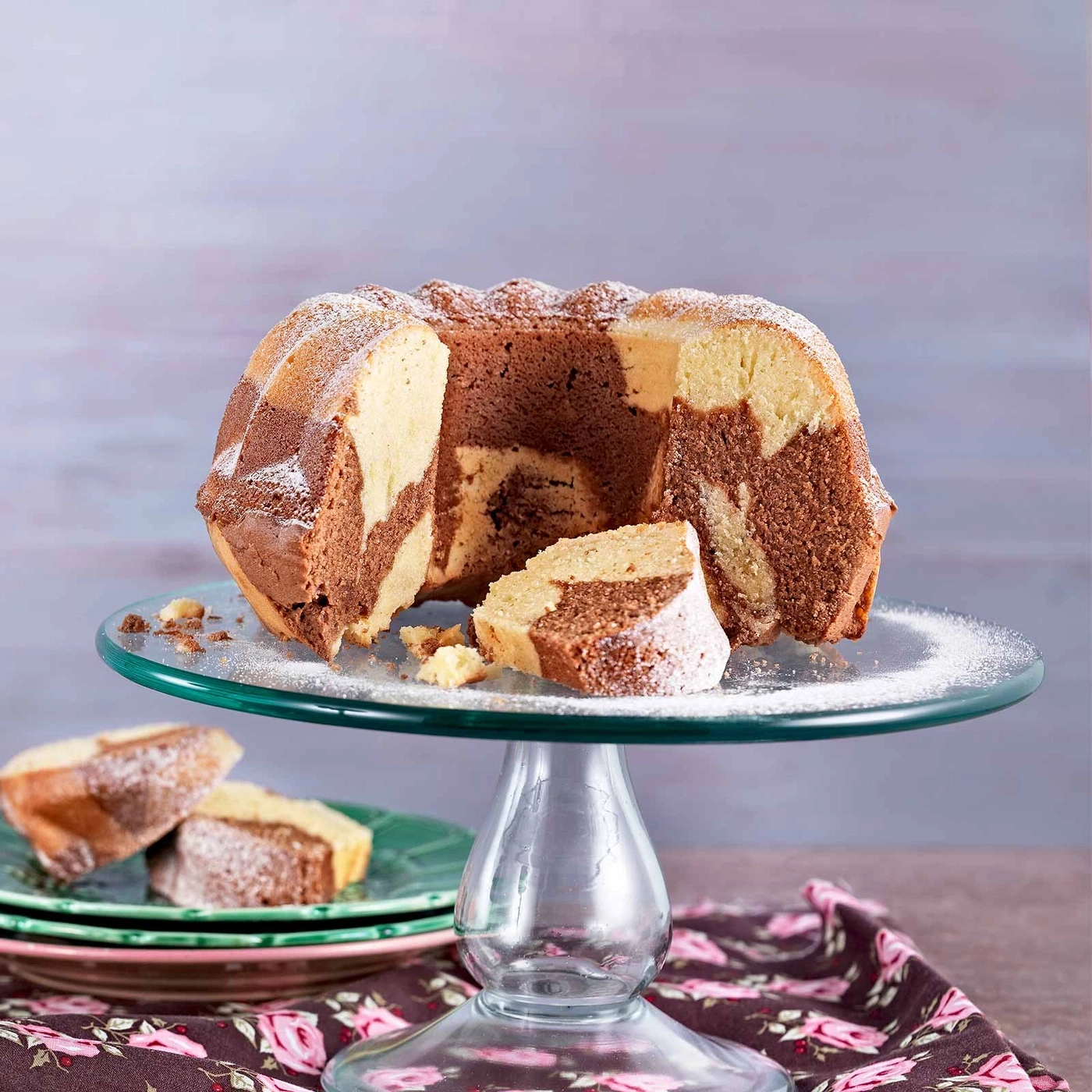 Marmorkuchen auf Glasplatte, mit Puderzucker bestäubt, angeschnitten, serviert mit Kuchenstücken.