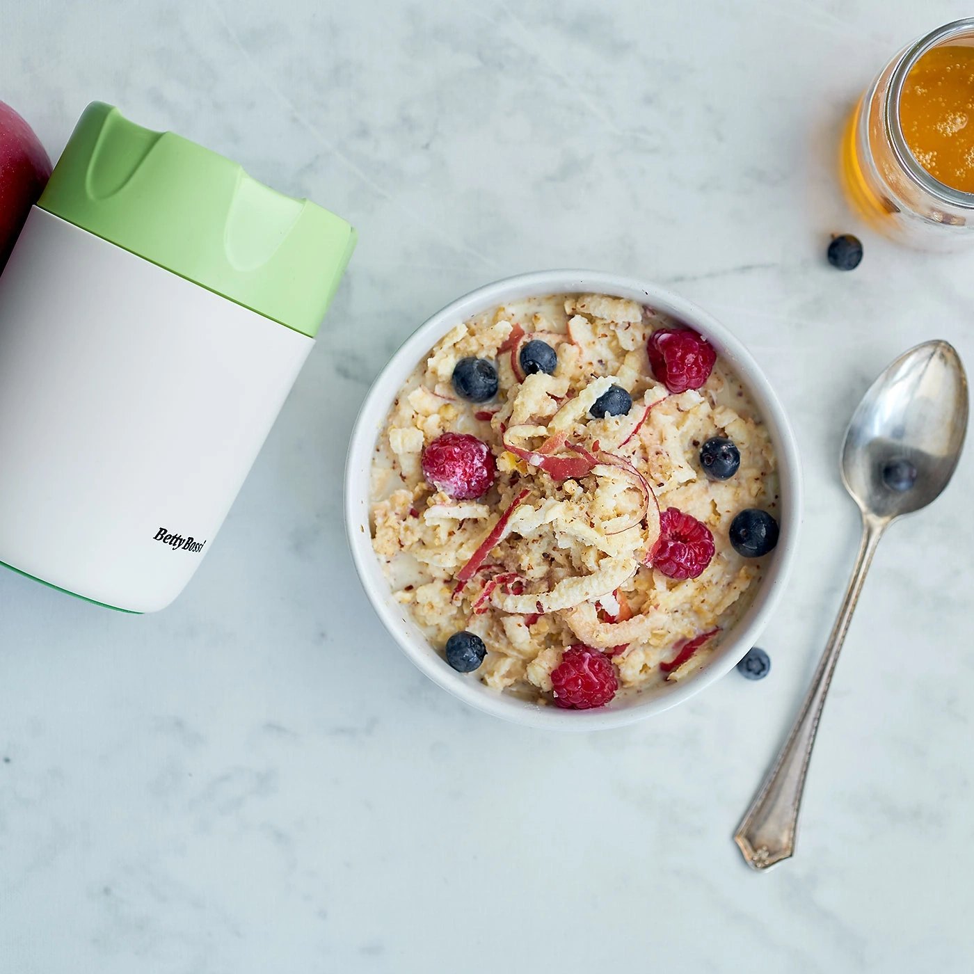 Schüssel mit Haferflocken, Beeren und Apfel, daneben Thermoskanne und Löffel