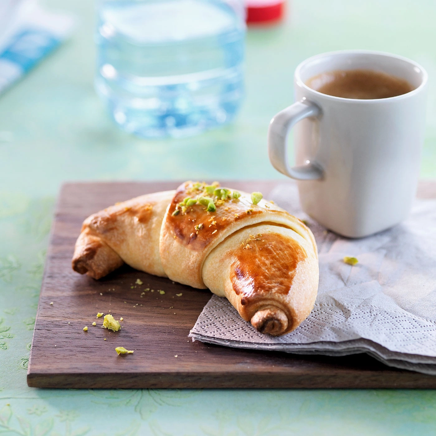Croissant mit Pistazien, Kaffee und Wasserflasche auf Holzbrett